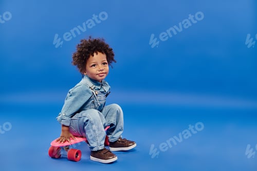 Preview: adorable african american toddler boy in denim clothes sitting on penny board on blue backdrop