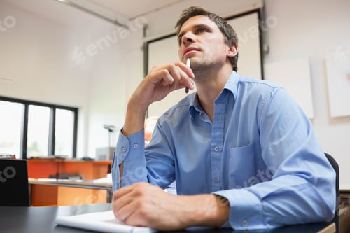Preview: Man sitting in meeting room at table holding pen near chin, viewing projection screen and notebook
