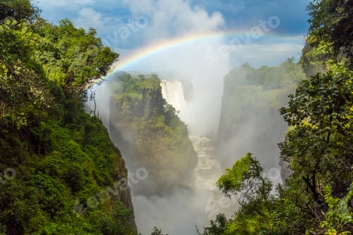 Preview: High angle view of waterfall cascading down overgrown rocks, single rainbow.