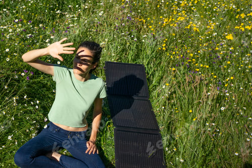 Preview: Woman sitting in wildflower meadow protecting her eyes from sunlight with portable solar panel