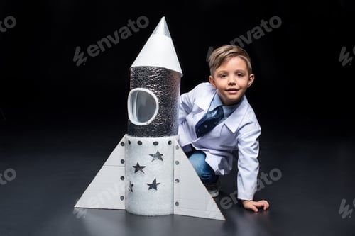 Preview: Cute little boy in white coat crouching near toy rocket and smiling at camera