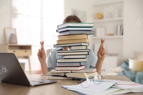 Preview: Student crossing fingers behind stack of books at table indoors. Hope for good exam result