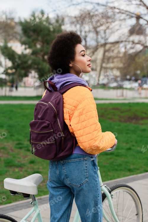 Preview: Side view of african american woman with backpack looking away near bicycle outdoors