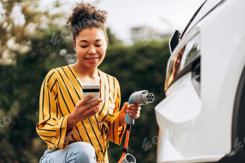 Preview: Happy African American woman using smartphone and charging car at vehicle charging station
