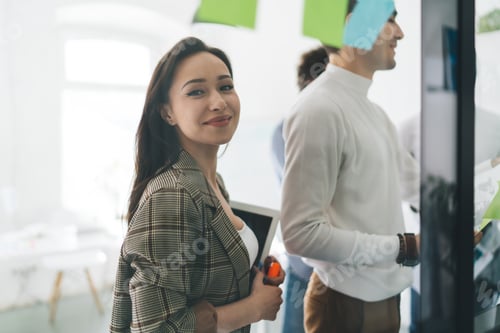 Preview: Young female designer standing in office near colleagues working on glass wall