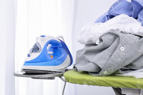 Vista previa: Modern electric iron and heap of clothes on board indoors. Laundry day