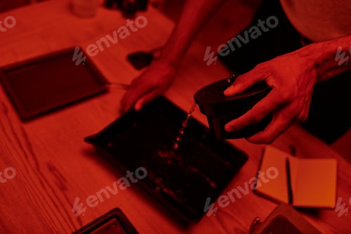 Preview: overhead view of photographer pours developer into tray for film processing in red darkroom