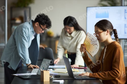 Preview: Young Asian Businesswoman Using Hand Fan While Collaborating with Colleagues