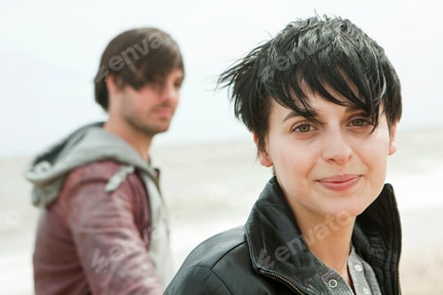 Preview: Woman with short hair smiling on a breezy beach day with a man in the background.