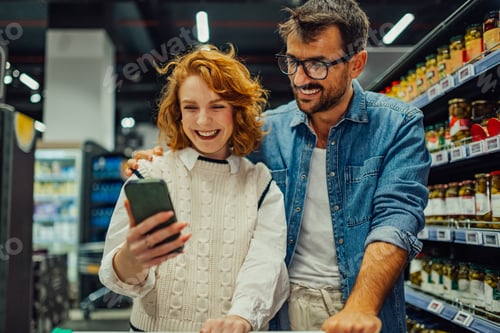 Preview: Happy couple using smartphone while shopping groceries in supermarket