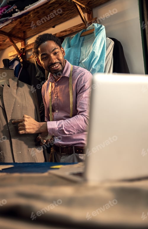 Preview: Male fashion tailor having video call with customer via laptop for the suit design in his workshop.
