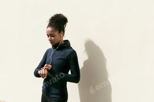 Preview: Young woman checking activity tracker on her wrist during outdoor exercise, standing at wall