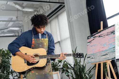 Preview: Young african american artist in apron playing acoustic guitar in workshop
