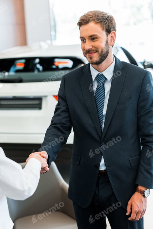 Preview: cropped view of woman shaking hands with happy car dealer in car showroom