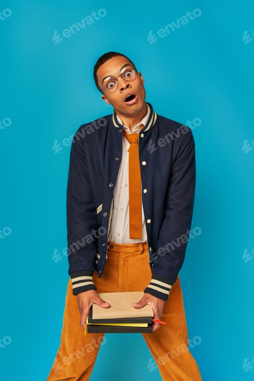 Preview: shocked african american student in stylish clothes holding notebooks and looking at camera on blue
