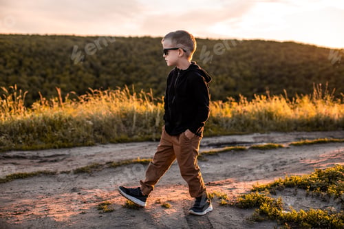 Preview: A boy in a black strong jacket and brown pants walks along a country road at sunset.