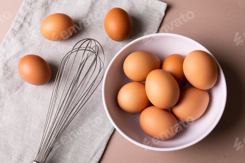 Preview: Directly above view of fresh brown eggs in bowl by wire whisk on napkin over table