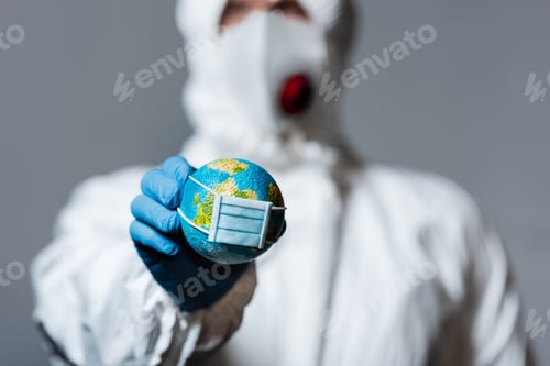 Preview: selective focus of man in hazmat suit holding small globe in medical mask isolated on grey