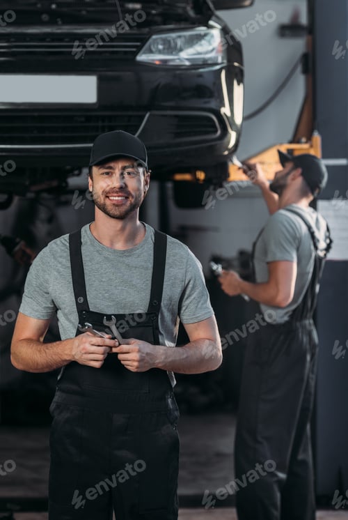 Preview: auto mechanic in overalls holding wrenches, while colleague working in workshop behind
