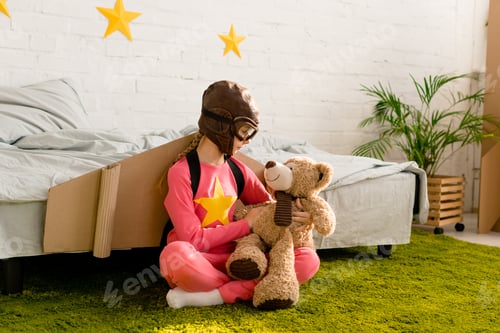 Preview: Child with cardboard wings sitting on carpet and looking at teddy bear