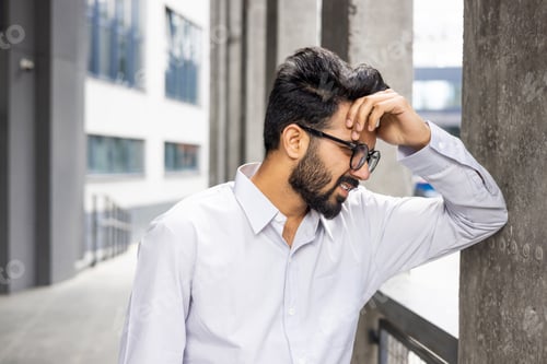 Preview: Tired young Indian man standing near a building, leaning against a wall and holding his head with