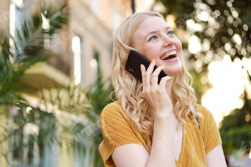 Preview: Portrait of pretty cheerful blond girl joyfully looking away talking on cellphone on city street