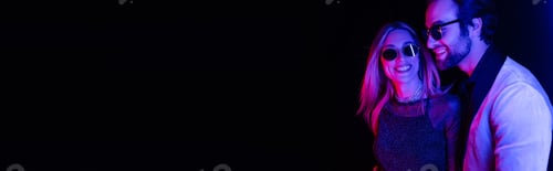 Preview: Smiling woman in sunglasses standing near stylish man isolated on black with colorful lighting,
