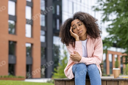 Preview: Pensive young woman sitting outdoors in urban setting feeling thoughtful and lost in her thoughts
