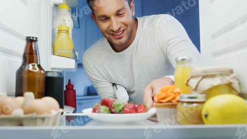 Preview: joyful man taking ripe strawberries from fridge with beverages, sauces, eggs and jars with food