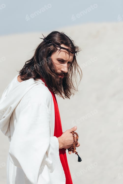Preview: side view of Jesus in robe and red sash holding wooden rosary in desert