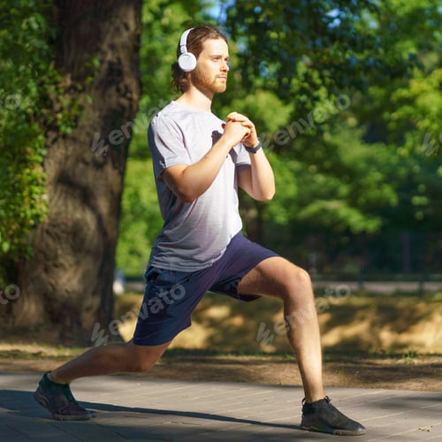 Preview: Young male sportsman in headphones doing sports exercises lunges outdoors