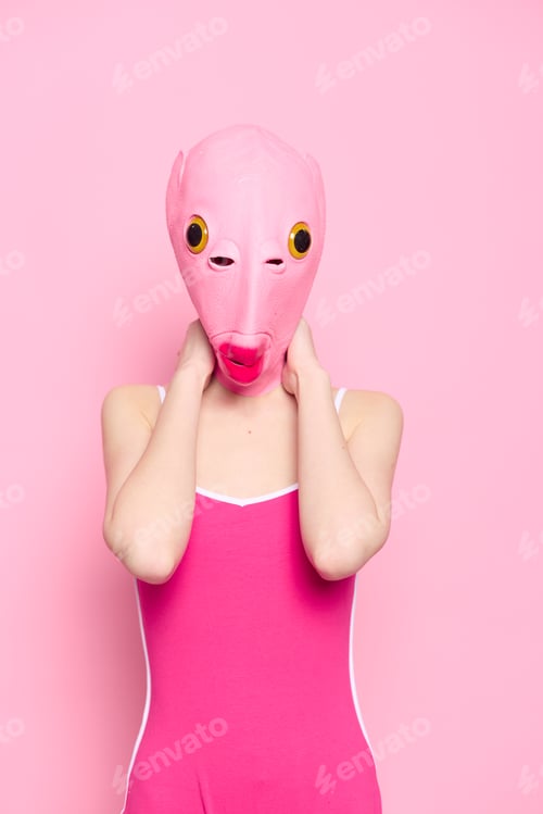 Preview: Woman standing in a pink fish mask on her head, a provocative crazy Halloween look, pink background