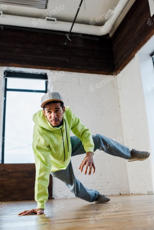 Preview: stylish and young african american man in cap breakdancing in dance studio