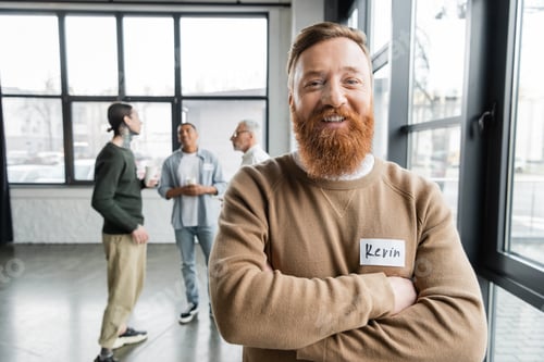 Preview: Cheerful bearded man crossing arms and looking at camera during alcoholics meeting in rehab center