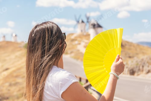 Preview: Blonde girl with a yellow fan in her hand fanning herself. Traditional windmills in the background.