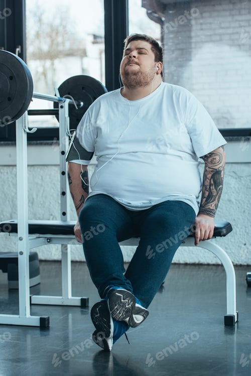 Preview: overweight tattooed man in earphones sitting on bench and listening music at gym