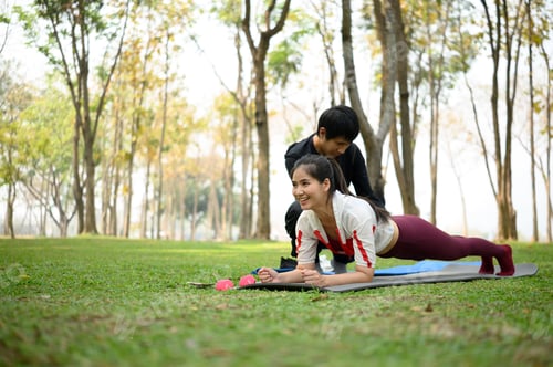 Preview: Strong young woman doing plank pose outdoors with trainer assistance