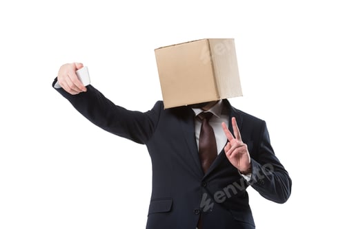 Preview: businessman with cardboard box on head and taking selfie with victory sign on smartphone, isolated