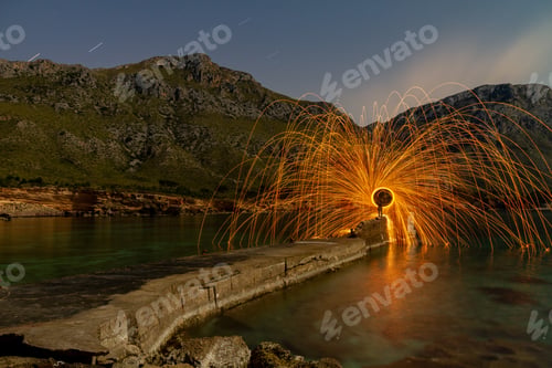 Preview: Person with burning circle on stones between water near hills at night