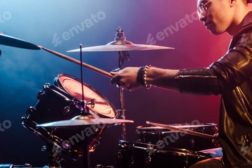 Preview: mixed race male musician in leather jacket playing drums during rock concert on stage with smoke and