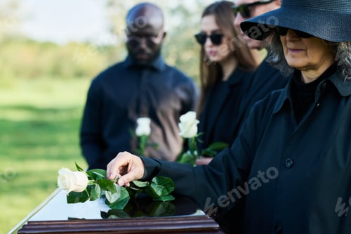 Preview: Mature grieving woman in mourning clothes putting white rose on coffin top