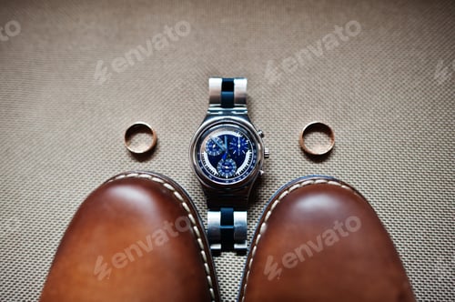 Preview: Close-up photo of groom's brown oxford shoes, a watch and two wedding rings.