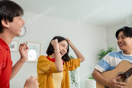 Preview: Diverse Asian friends playing guitar and dancing in living room at home.