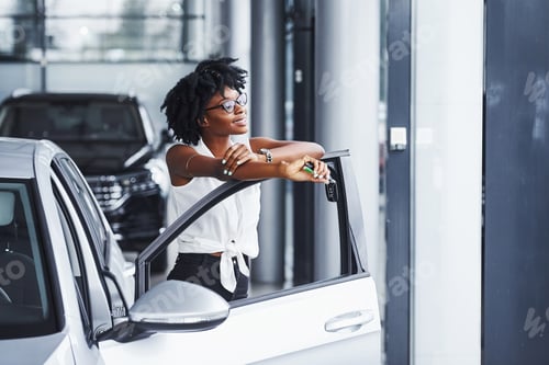 Preview: Young african american woman in glasses stands in the car salon near vehicle with keys in hands