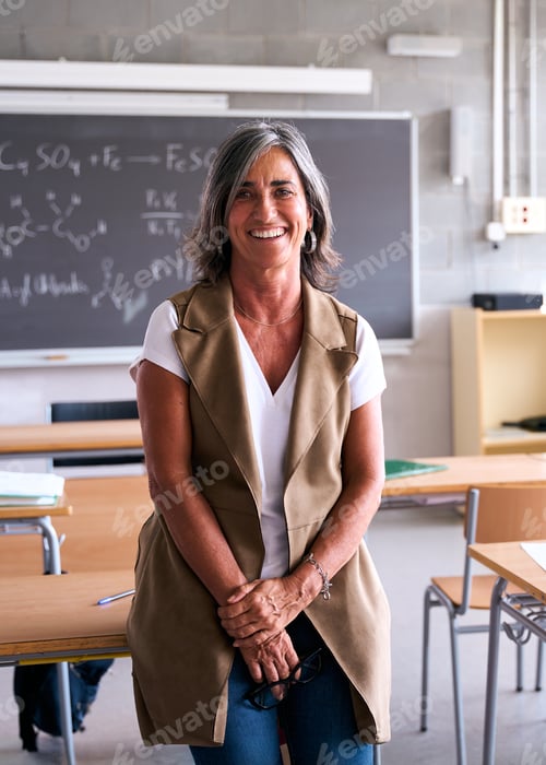 Visualização: Professora de meia idade do ensino médio olhando sorrindo para a câmera com as mãos juntas na sala de aula.