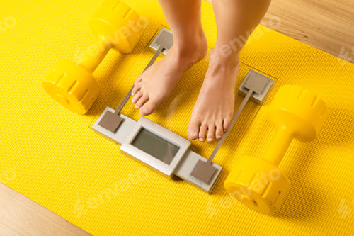 Preview: Female feet on the modern weighing scale with a yellow dumbbells and fitness mat