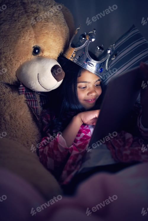 Preview: Cropped shot of a little girl using a digital tablet while lying in bed with her teddy at night