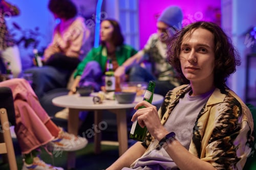 Preview: Young man with bottle of beer in hand looking at camera at home party