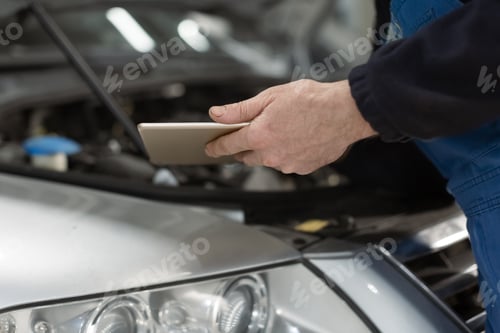Preview: Hands of mature white auto mechanic holding tablet PC, blurred car on the background