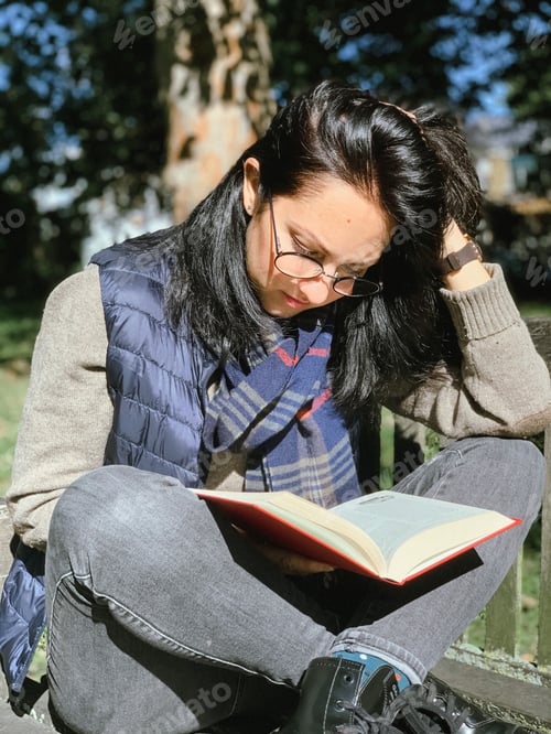 Preview: Young candid woman in casual autumnal clothes siting on a wooden bench in park and reading red book
